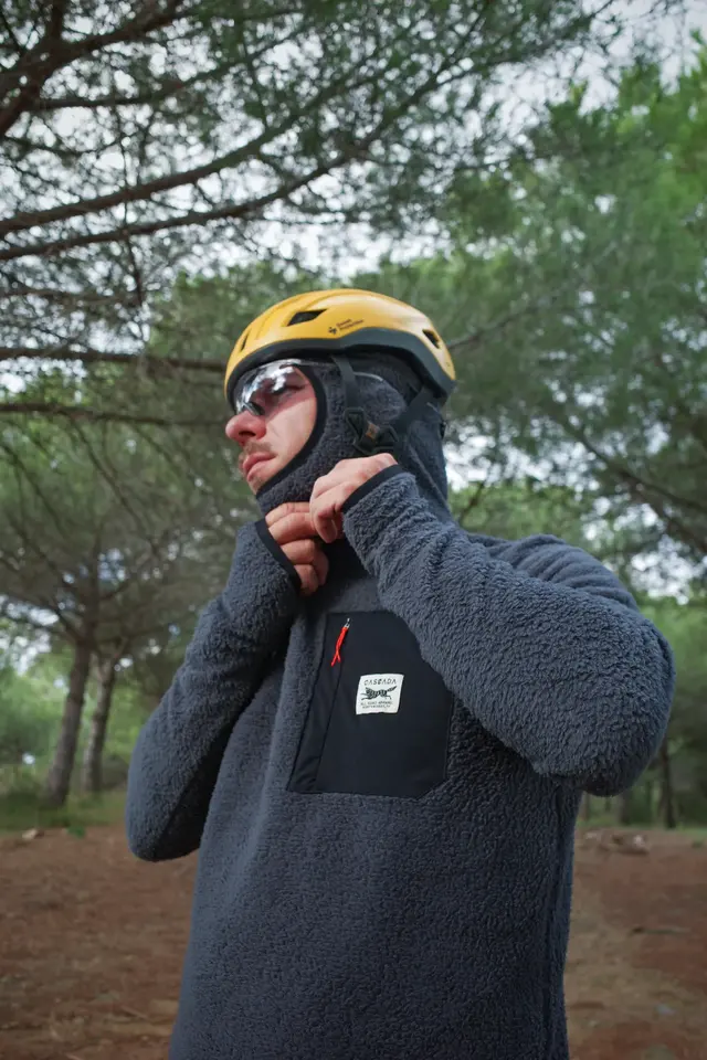 A person in a grey jacket and yellow helmet standing among pines