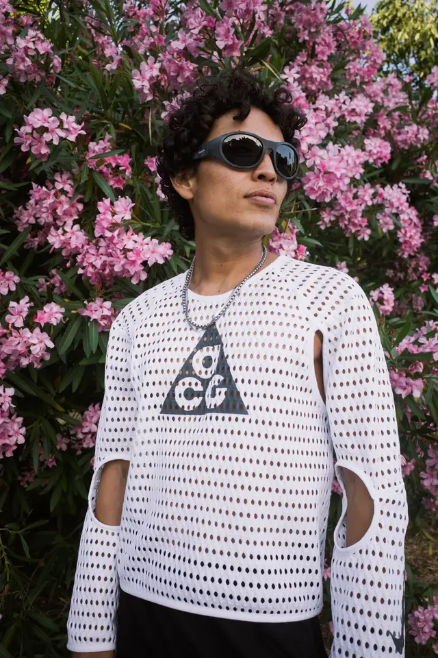 A person in sunglasses and a white open-knit shirt, standing among pink flowers